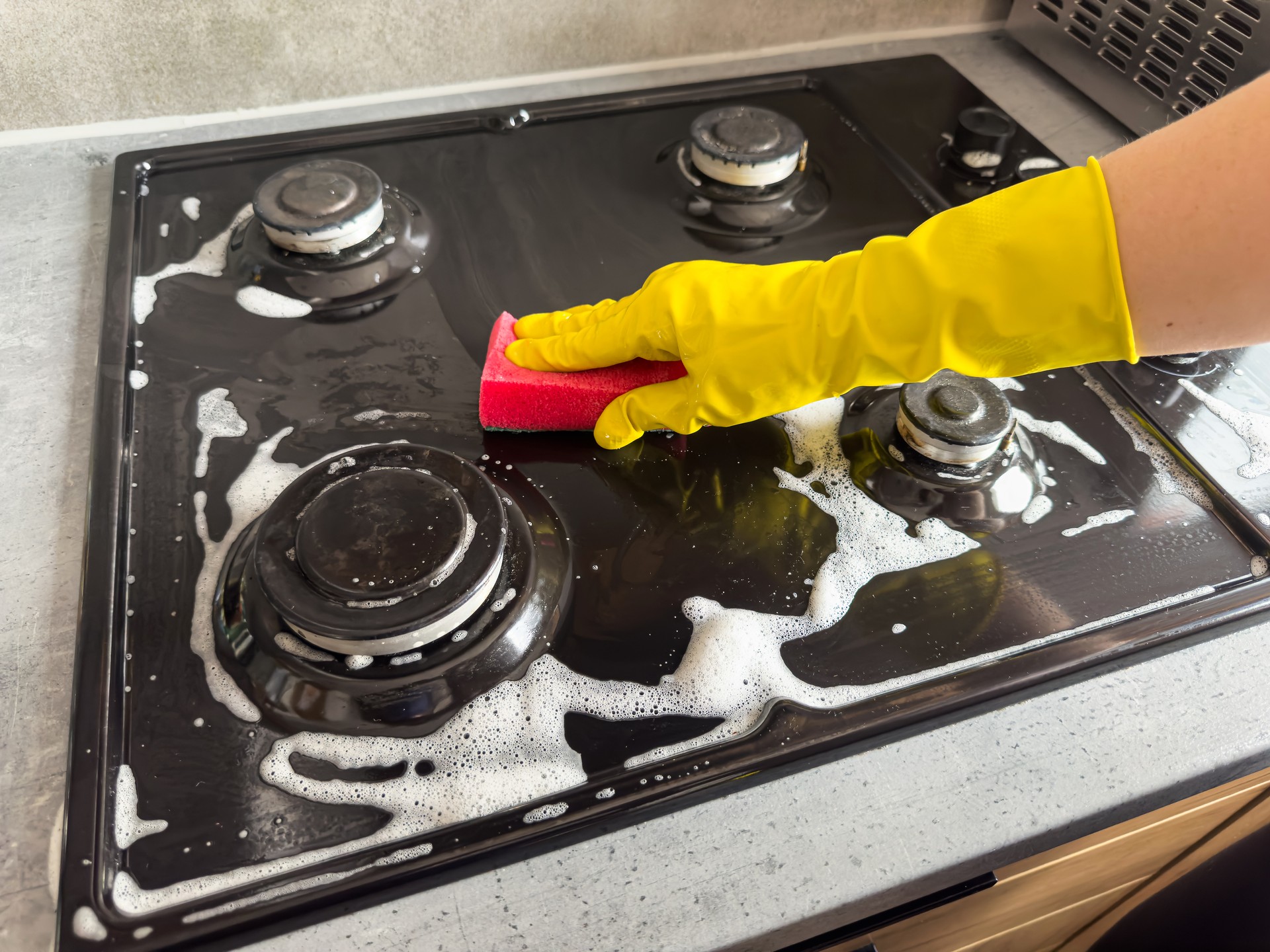 female hand in gloves with rag sponge a foam cleaning gas stove at home kitchen